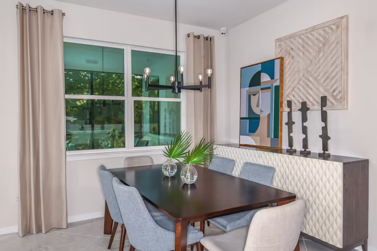 A modern dining room with a dark wood table, six upholstered chairs, two small vases with palm leaves, abstract artwork, decorative sculptures, beige curtains, and a geometric chandelier.