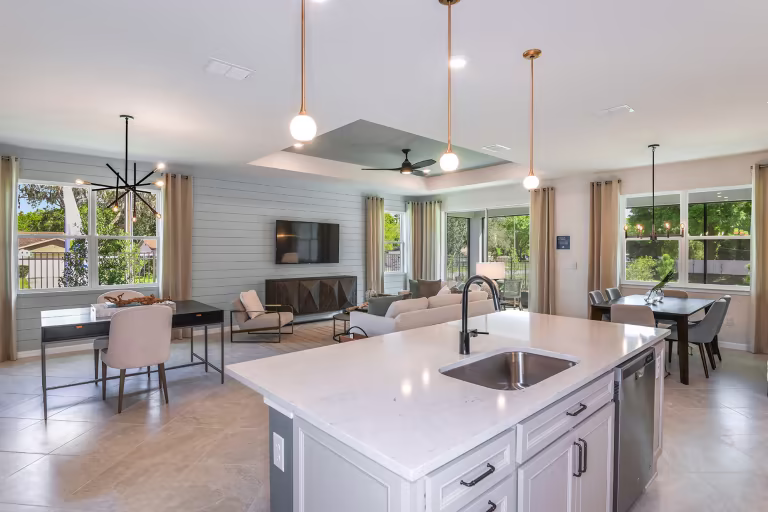 A modern, open-concept kitchen and living area with white countertops, pendant lights, a large island with a sink, dining table, sofa, TV, and large windows letting in natural light.