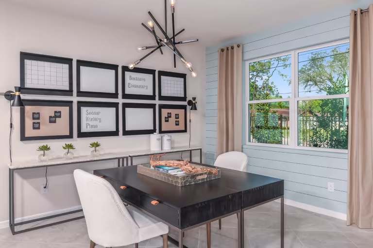 A modern home office with a black desk, two white chairs, framed wall art, succulents, a large window with beige curtains, and a unique black chandelier. The space has light walls and natural lighting.