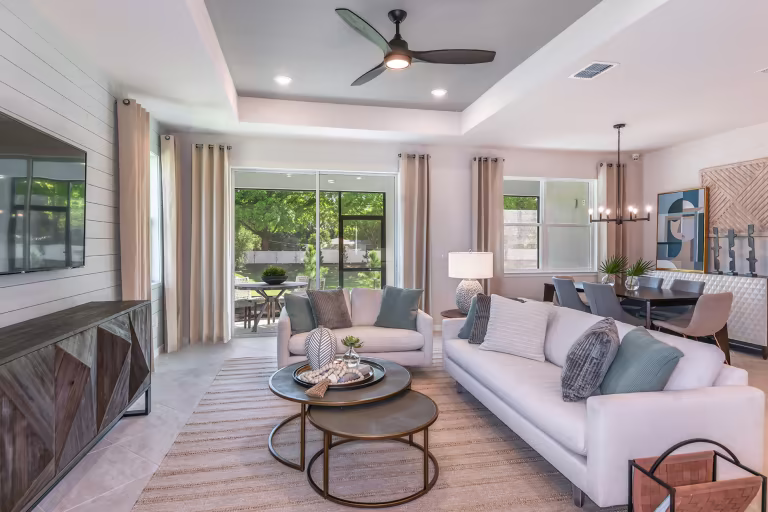 Bright, modern living room with two white sofas, round coffee tables, large windows with beige curtains, a ceiling fan, wall-mounted TV, and an adjacent dining area with a table and chandelier.