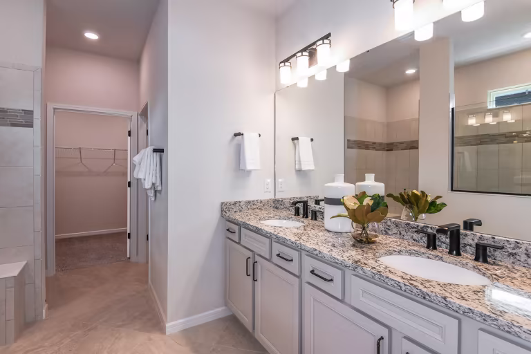 Modern bathroom with double sinks, granite countertop, large mirror, black fixtures, white cabinetry, decorative vases, towel racks, and an adjoining walk-in closet with hanging space visible through an open doorway.