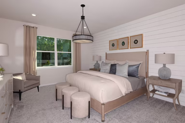 A modern bedroom with a beige upholstered bed, gray bedding, three round ottomans at the foot, two nightstands with lamps, a dresser, an armchair, and a window with curtains letting in natural light.