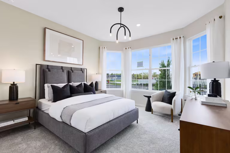Bright, modern bedroom with a gray upholstered bed, white bedding, black pillows, two bedside tables with lamps, a chair by large windows, white curtains, and a wooden dresser. Minimalist decor with neutral tones.