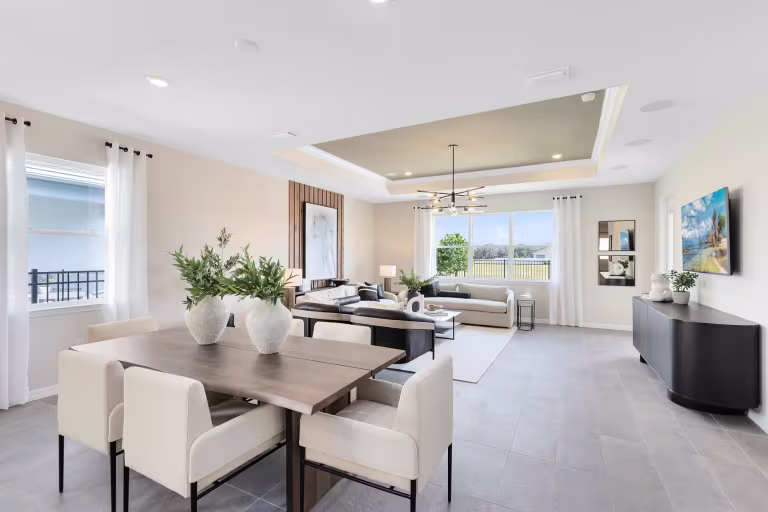 Modern open-plan living and dining area with large windows, neutral tones, a wooden dining table with white chairs, two sofas, decorative plants, and contemporary artwork on the walls.