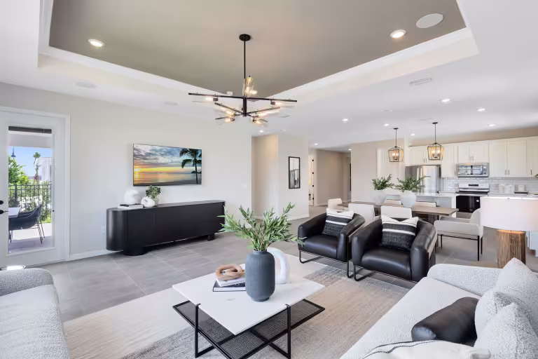 Modern open-concept living room and kitchen with neutral decor, contemporary furniture, a large wall-mounted TV, stylish chandelier, and natural light from glass doors leading to a balcony.