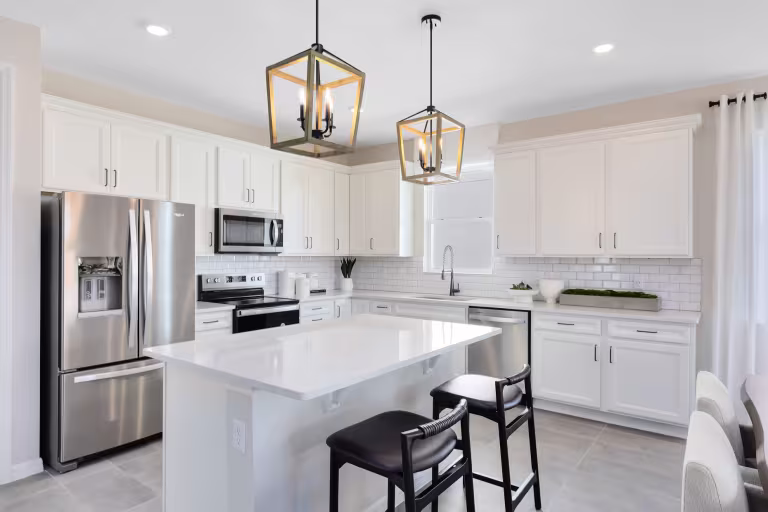 Modern kitchen with white cabinets, stainless steel appliances, a large island with two black stools, subway tile backsplash, and two geometric pendant lights hanging from the ceiling.