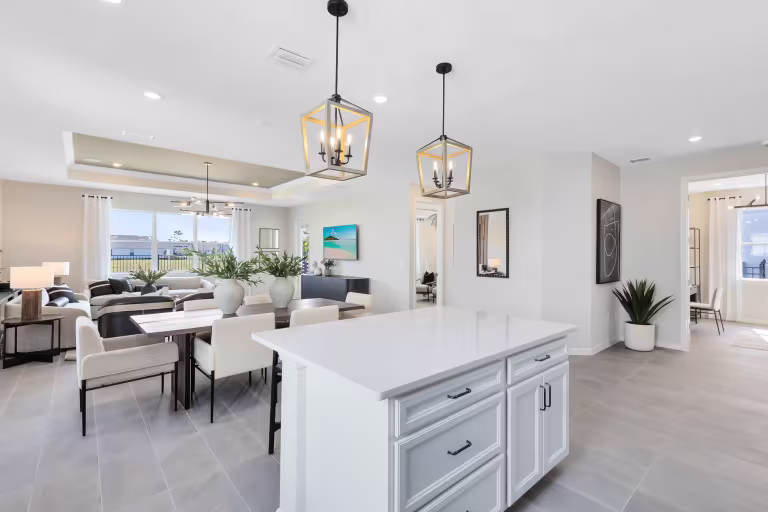 Modern open-concept kitchen and living room with white cabinets, an island, pendant lights, dining table, sofas, large windows, and neutral decor. Bright, spacious, and contemporary interior design.