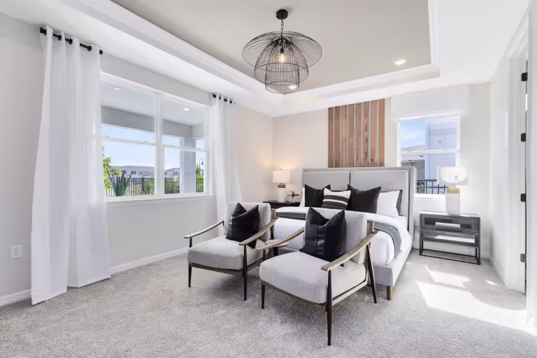 Modern bedroom with a large bed, two armchairs, and decorative pillows. The room features light walls, large windows with white curtains, a carpeted floor, and a contemporary ceiling light fixture.
