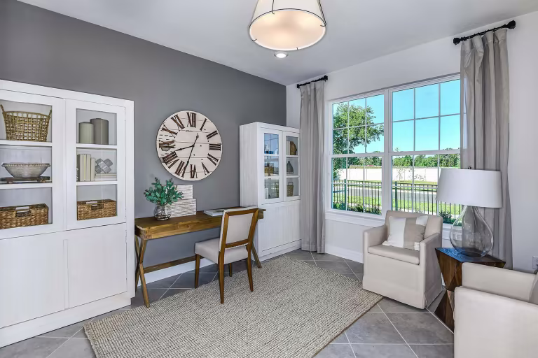 A bright home office features a wooden desk and chair, white shelves with baskets and books, a large clock on a gray accent wall, an armchair, a lamp, and a window with beige curtains overlooking greenery.