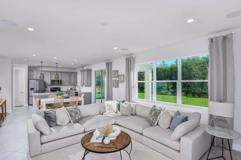Bright, modern open-concept living room and kitchen with a large beige sectional sofa, round coffee table, dining area, grey cabinets, pendant lights, and large window overlooking a lush green backyard.