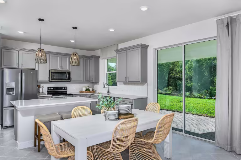 Modern kitchen and dining area with gray cabinets, stainless steel appliances, a white island with barstools, a white dining table with wicker chairs, and large sliding glass doors overlooking a green backyard.