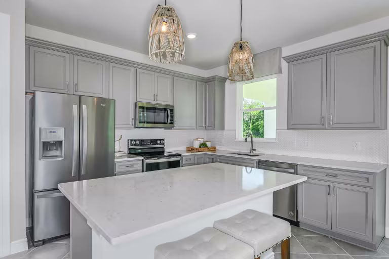 Modern kitchen with gray cabinets, stainless steel appliances, a large white island with two tufted stools, pendant lights overhead, and a window above the sink letting in natural light.