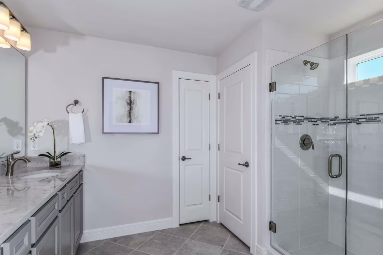 Modern bathroom with a glass shower enclosure, marble countertop, double sink, grey cabinets, orchid plant, framed wall art, towel ring, and two white doors. The space is bright with natural light.