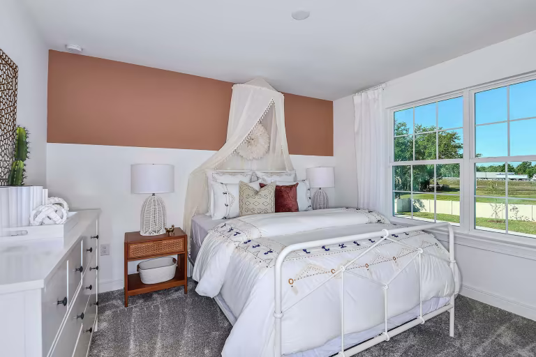 Bright bedroom with a white metal bed, neutral bedding, decorative pillows, two bedside tables with lamps, a dresser, and a large window letting in natural light. A canopy hangs above the bed on a beige accent wall.