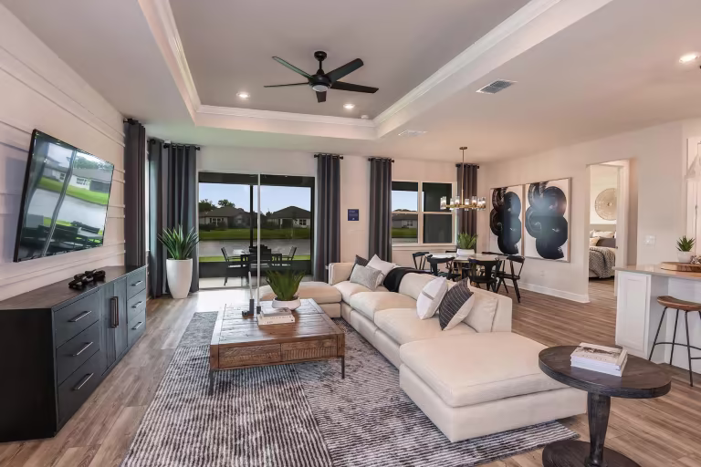 Modern living room with a large sectional sofa, coffee table, TV, and floor-to-ceiling windows. Dining area and bedroom are visible, with wood flooring and neutral decor throughout.
