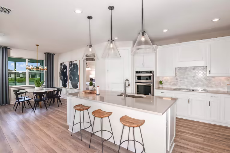 Modern kitchen with white cabinets, a large island with three wooden stools, pendant lights, stainless steel appliances, and an open dining area with a window and contemporary artwork on the wall.