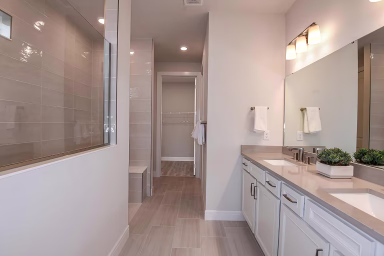 Modern bathroom with double sink vanity, large mirror, wall-mounted lights, glass-enclosed shower with bench, beige tile floors, and neutral-colored walls. Small green plants decorate the countertop.