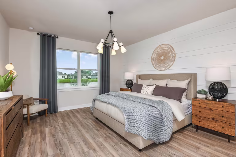 Modern bedroom with wood floors, a large bed with gray and white bedding, two wooden nightstands with lamps, a dresser with plants, a gray armchair, a large window with curtains, and a decorative wall piece above the bed.