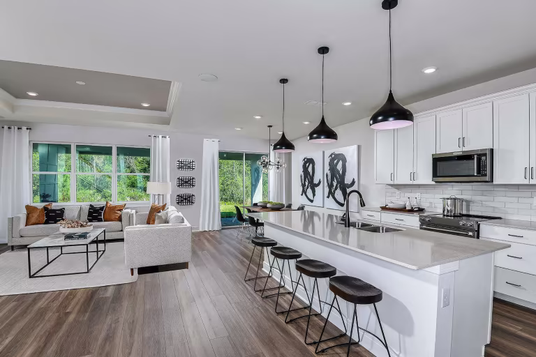 Modern open-concept kitchen and living area with white cabinets, stainless steel appliances, a large island with stools, pendant lighting, and a cozy seating area near large windows overlooking greenery.