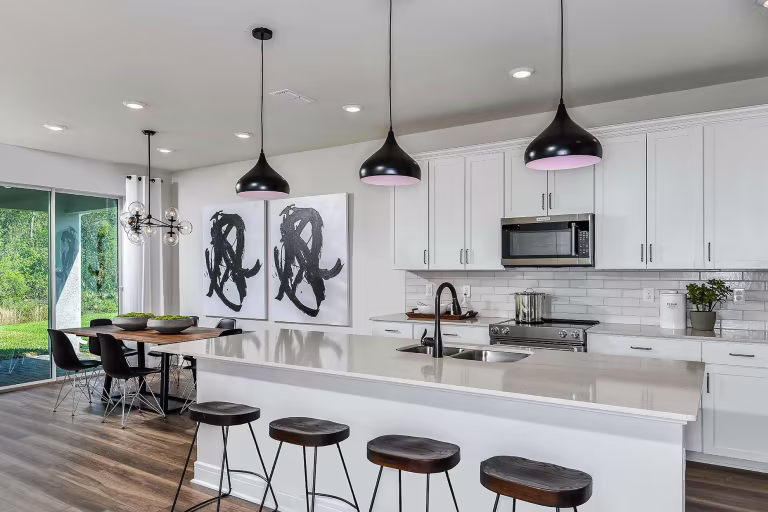 Modern kitchen with a large white island, four dark wooden stools, pendant lights, white cabinets, stainless steel appliances, and a dining area with abstract art and large windows overlooking greenery.