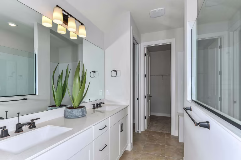 Modern bathroom with white cabinets, a large mirror, double sink, black fixtures, three-light vanity, two tall potted plants on the counter, and a view into a closet through an open door.