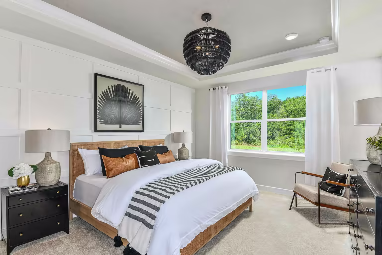 Modern bedroom with a large bed featuring white and black bedding, two nightstands with lamps, an armchair, a dresser, a black pendant light, artwork above the bed, and a large window letting in natural light.
