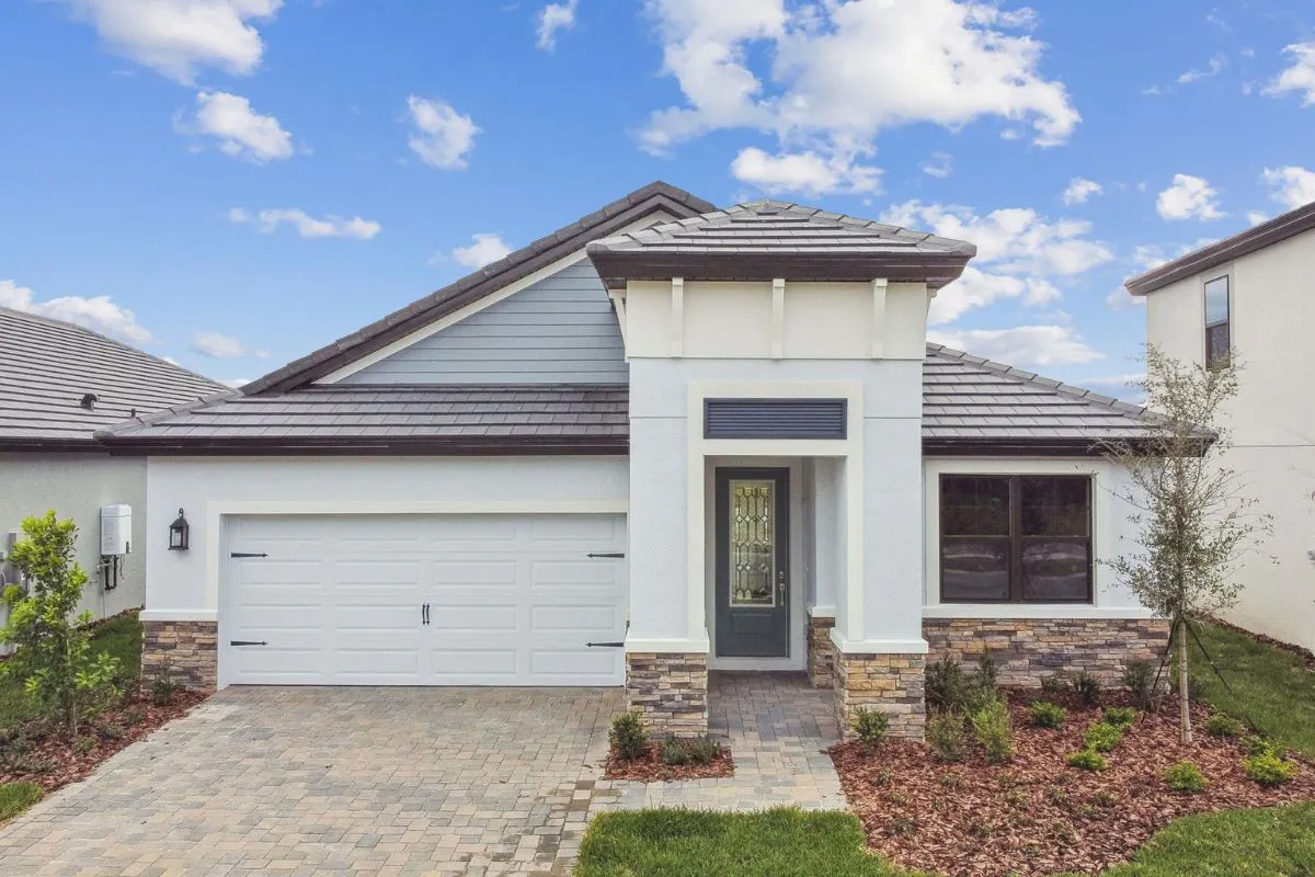Single-story modern house with white walls, grey roof, large front-facing garage, and a glass-panel front door. The yard has neatly landscaped plants, shrubs, and a stone walkway under a partly cloudy sky.