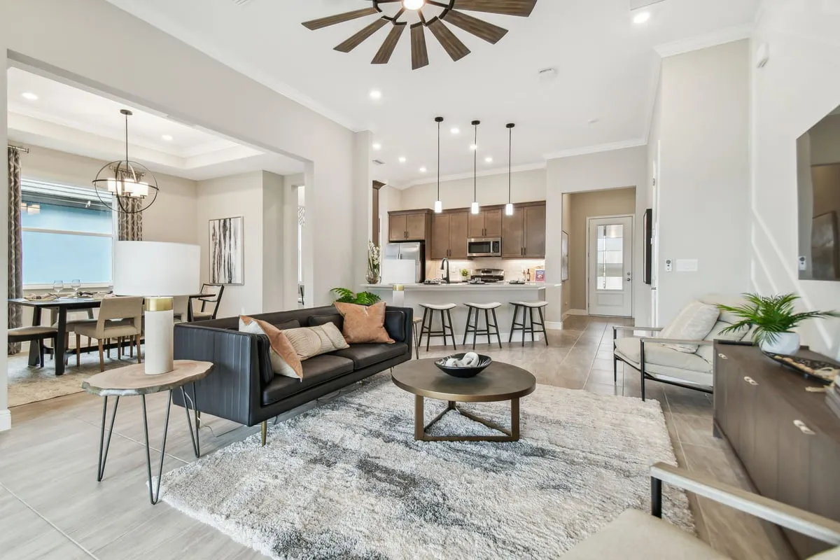 Modern open-concept living room with gray sofa, round coffee table, rug, and armchairs, leading to a kitchen with barstools and pendant lights, and a dining area with a large window and chandelier.