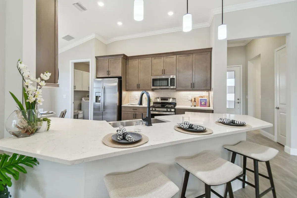 Modern kitchen with a white island countertop, three stools, stainless steel appliances, brown cabinets, a built-in microwave, and place settings with napkins. A vase with flowers sits on the counter. Bright lighting overhead.