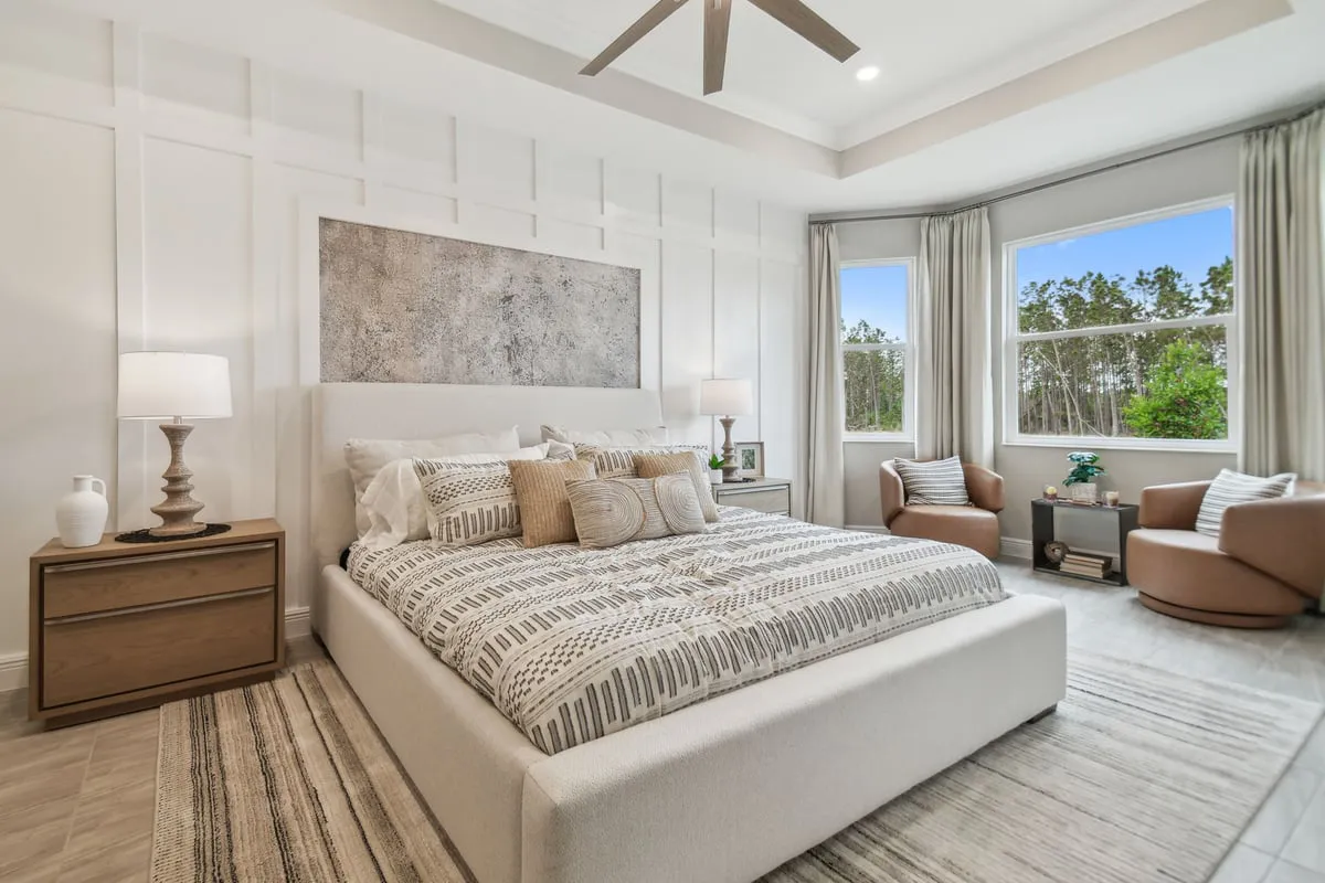 Modern bedroom with a large upholstered bed, patterned bedding, two bedside tables with lamps, two armchairs by tall windows, light curtains, and a view of trees outside. Neutral colors and soft lighting create a cozy feel.