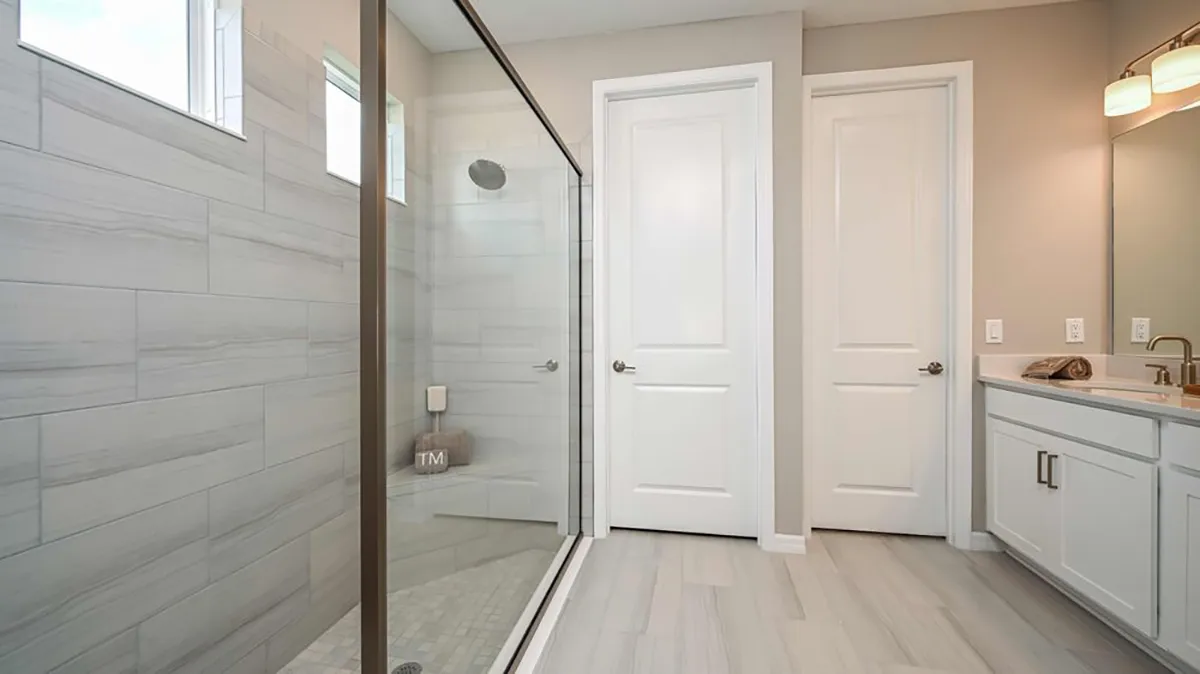 Modern bathroom with a large glass-enclosed shower, gray tile walls, two white doors, a white vanity with a sink, mirror, and light fixture above. Neutral color palette with clean lines and minimal decor.