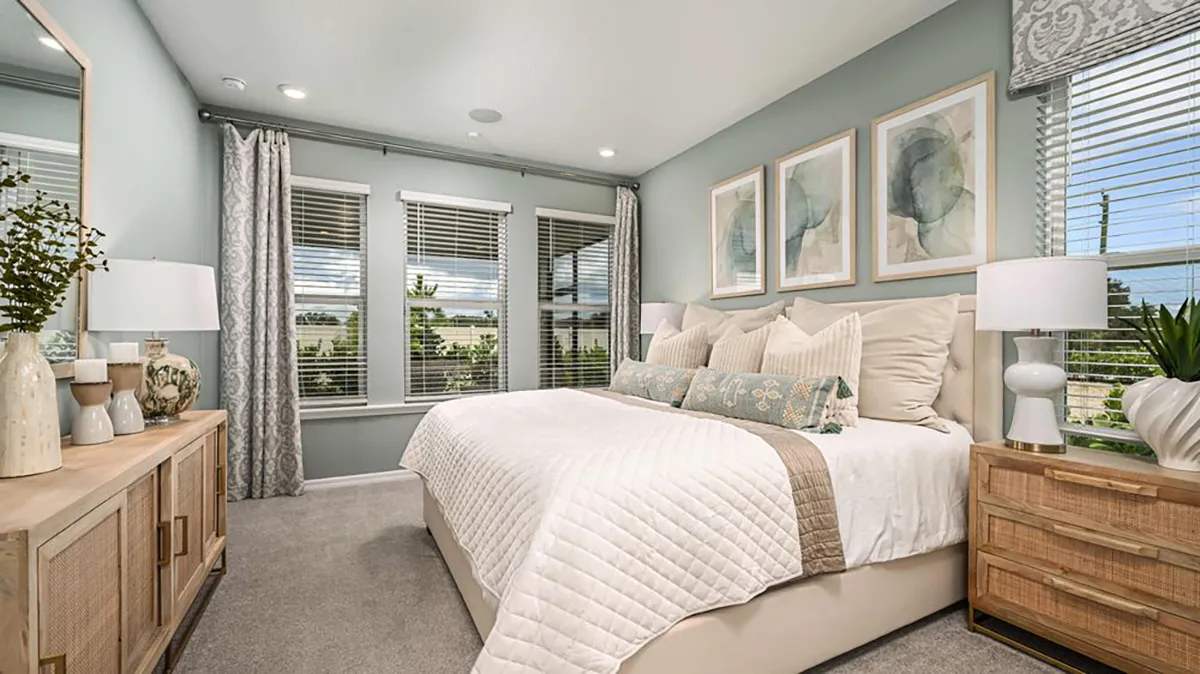 Modern bedroom with a large bed, white bedding, wooden nightstands with lamps, abstract art on the wall, a dresser with decorative items, and large windows with gray curtains letting in natural light.