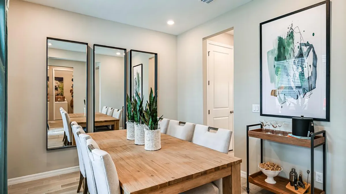A modern dining room with a wooden table, eight white cushioned chairs, three tall mirrors on one wall, a large abstract artwork, potted plants, and a small side cart with decor and snacks.