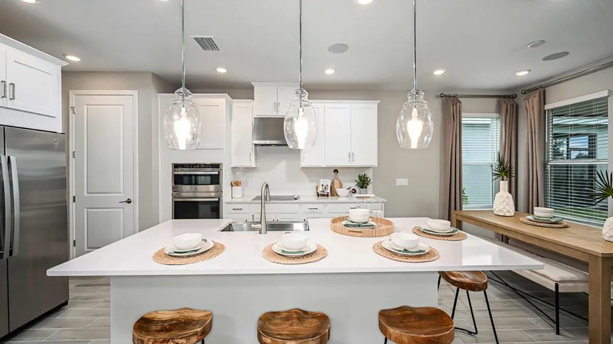 Modern kitchen with white cabinets, stainless steel appliances, island with four place settings, pendant lights, wooden stools, and a window-side dining table decorated with vases and greenery.