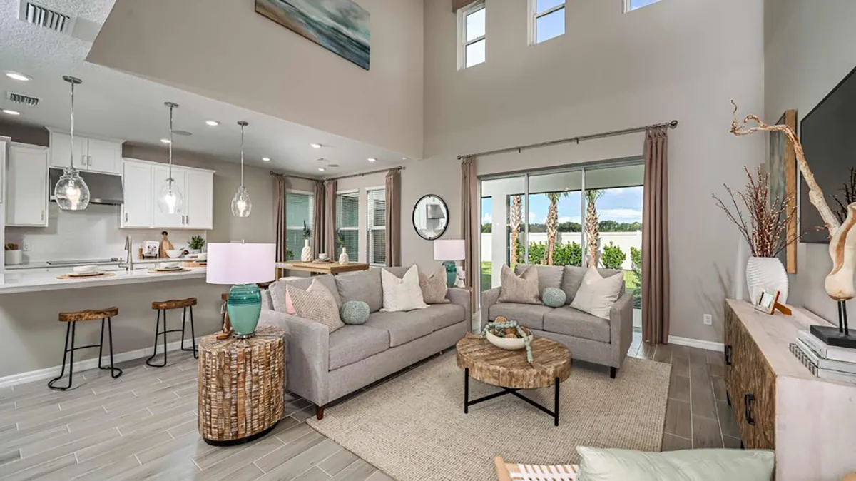 Modern open-concept living room and kitchen with neutral tones, two gray sofas, a round wooden coffee table, and large windows offering a scenic outdoor view. Kitchen features white cabinets and barstool seating.