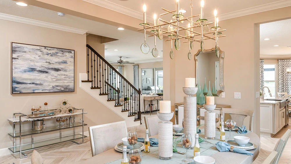 A modern dining room with a glass table set for six, white chairs, tall decorative candles, a large chandelier, and neutral-toned walls. A staircase and open kitchen are visible in the background.