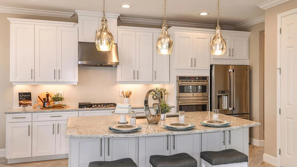 Modern kitchen with white cabinets, stainless steel appliances, marble island with three place settings, pendant lights above, and three gray cushioned stools tucked under the island.