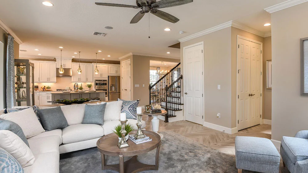 Modern living room with a white sectional sofa, gray ottoman, round wooden coffee table, decorative plants, and neutral walls. Open layout leads to a kitchen with pendant lights and a staircase with black railings.