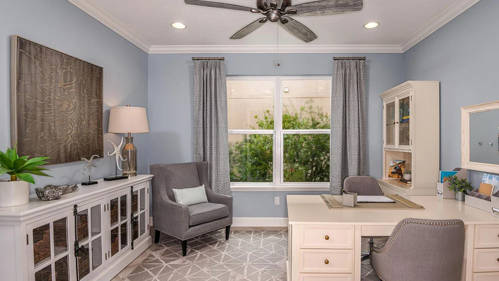 A home office with a white desk, gray upholstered chairs, built-in shelves, a table lamp, potted plants, and large windows with gray curtains letting in natural light. A ceiling fan is above, and the walls are painted light blue.
