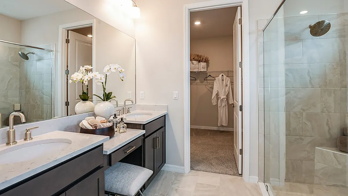Modern bathroom with double sinks, marble countertops, large glass shower, and light-colored tile floors. White orchids, a bench, and toiletries decorate the vanity. An open door reveals a walk-in closet with robes.