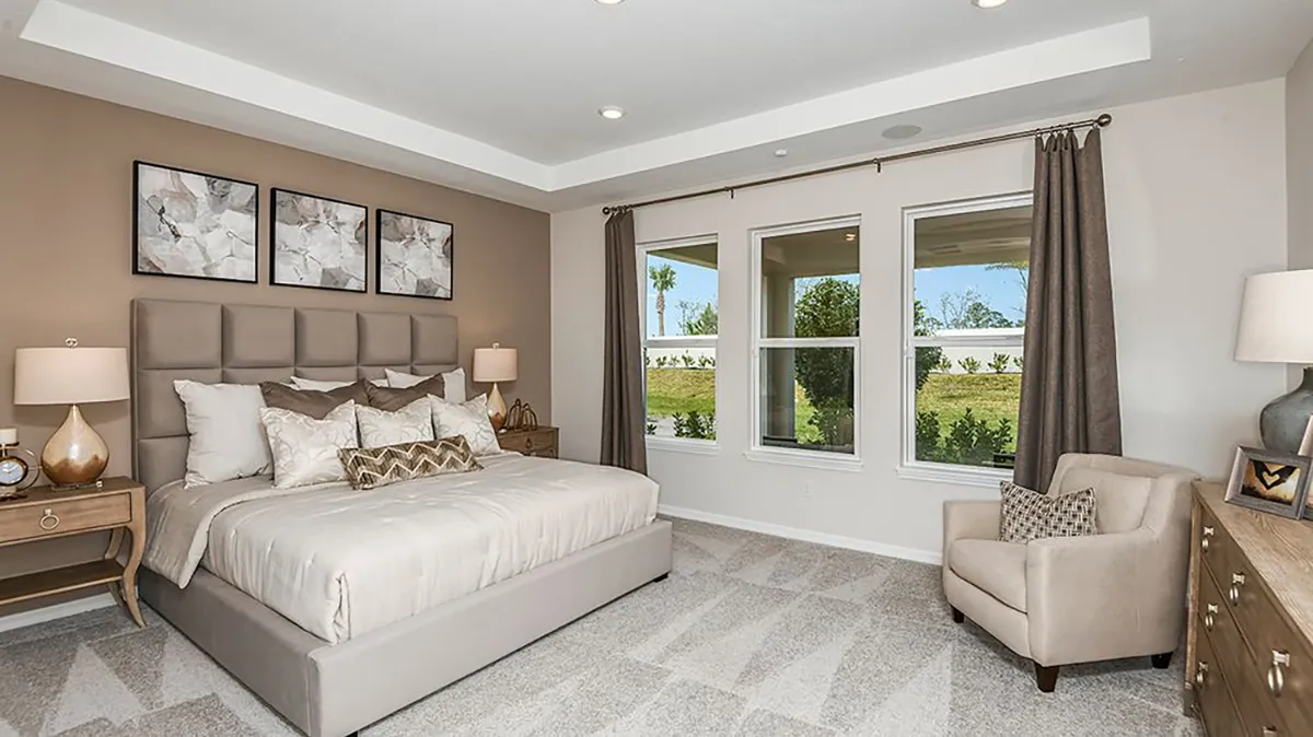 Modern bedroom with a large bed, neutral decor, abstract art above the headboard, two nightstands with lamps, a cushioned armchair, a dresser, and large windows with brown curtains overlooking greenery.