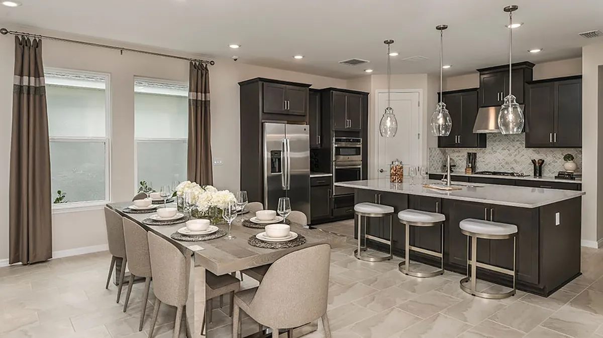 Modern kitchen and dining area with dark cabinets, stainless steel appliances, a center island with three stools, and a dining table set for six with neutral chairs, all under recessed and pendant lighting.