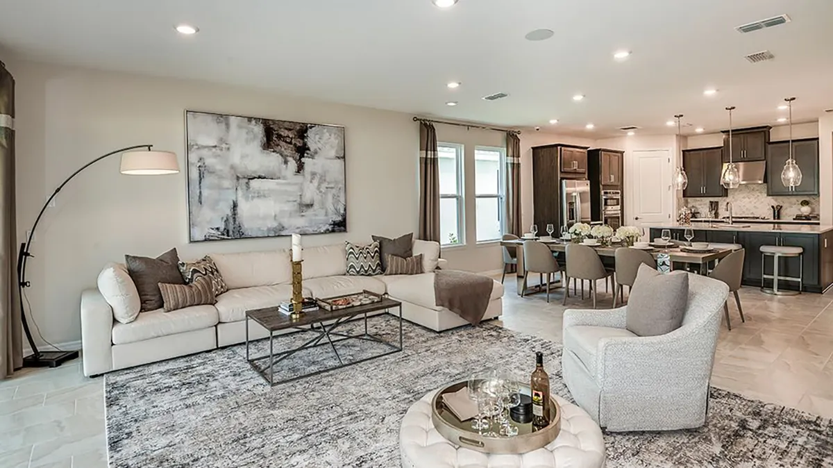 Modern open-concept living room and kitchen with neutral tones, a large sectional sofa, abstract wall art, a round ottoman, dining area, and pendant lights above a kitchen island.