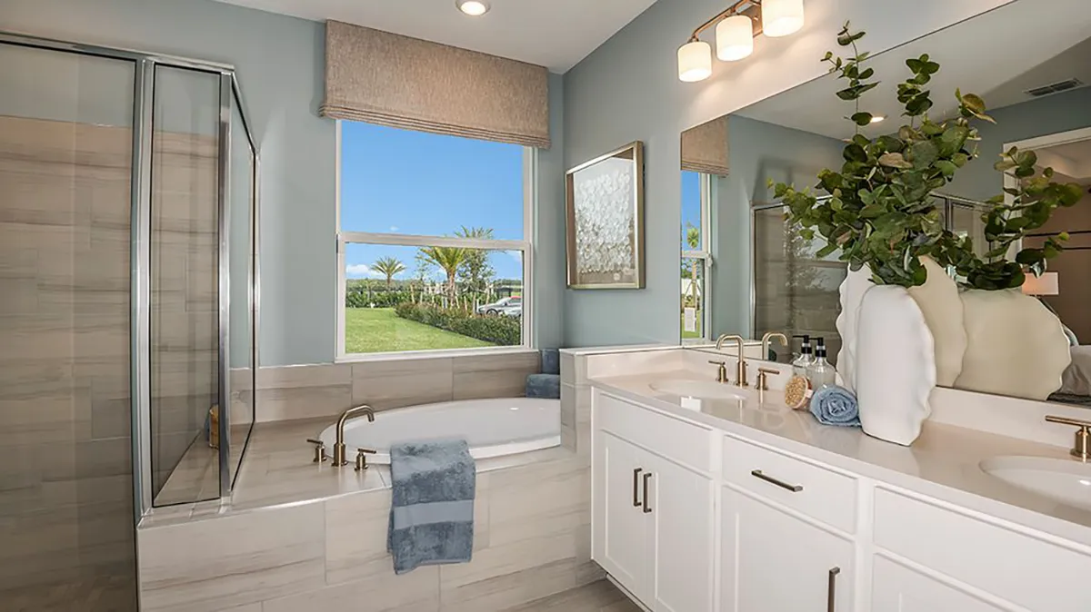 Modern bathroom with a glass shower, soaking tub, large window with a view of a grassy yard, double vanity with white cabinets, gold fixtures, and a decorative vase with greenery on the counter.