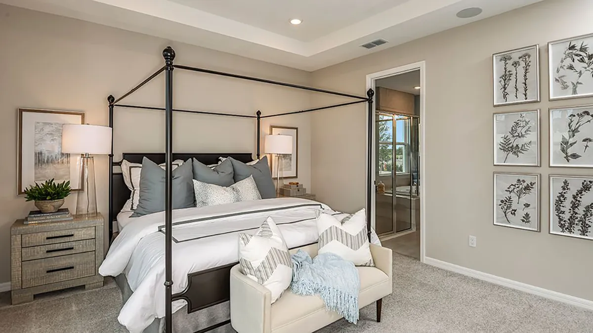 A modern bedroom with a black four-poster bed, gray and white bedding, a small bench at the foot, two nightstands with lamps, and framed botanical prints on the wall. An open doorway leads to an en suite bathroom.