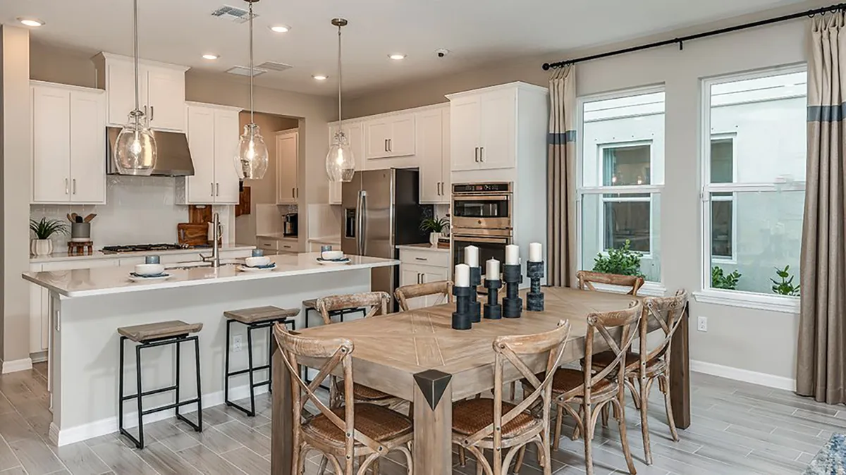 Modern kitchen and dining area with white cabinets, stainless steel appliances, a large island with stools, a wooden dining table with eight chairs, and decorative candles. Large windows let in natural light.