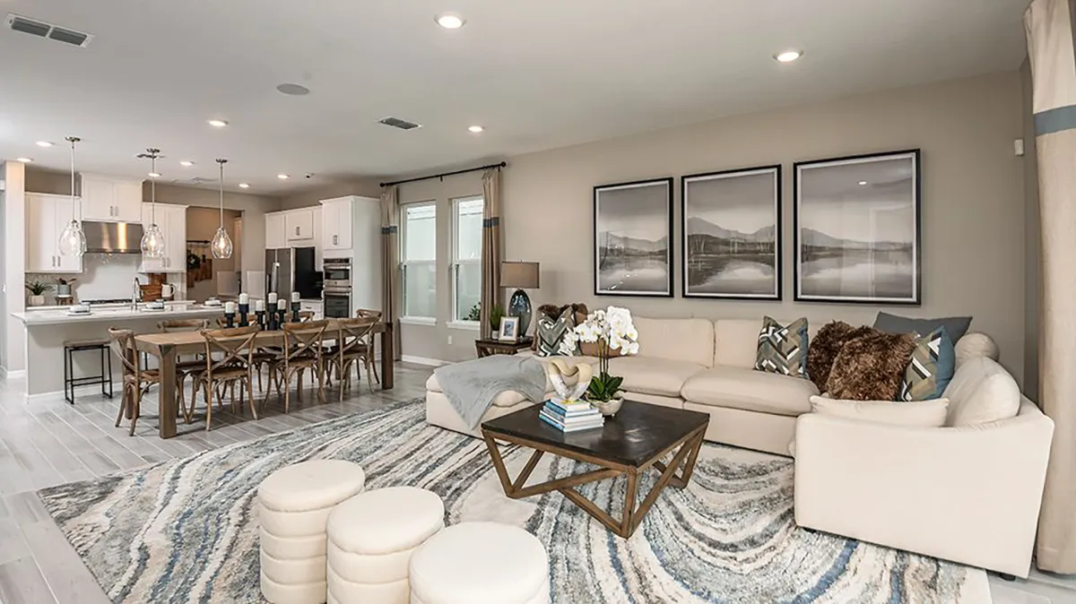 Modern open-concept living room and kitchen with beige sectional sofa, abstract rug, wooden dining table, and white cabinetry. Large windows and wall art add brightness and style to the space.