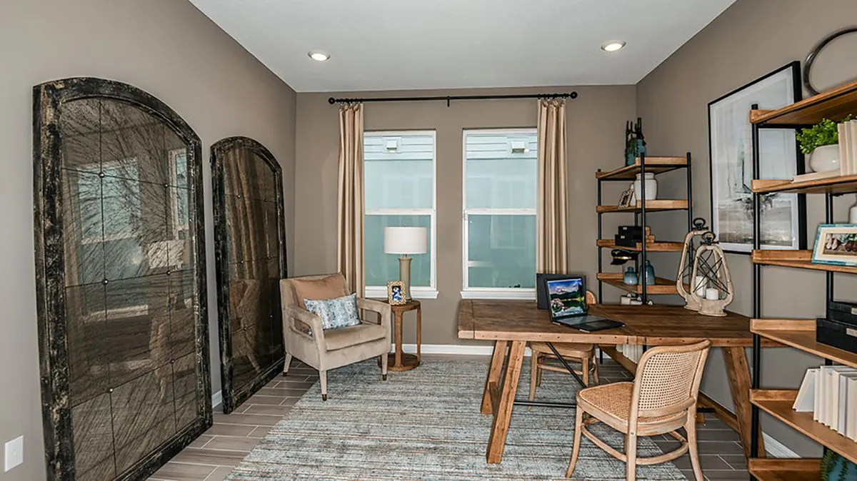 A modern home office with a wooden desk, rattan chair, beige armchair, tall shelving unit, and large windows with light curtains. Neutral walls and a patterned area rug create a cozy, organized workspace.
