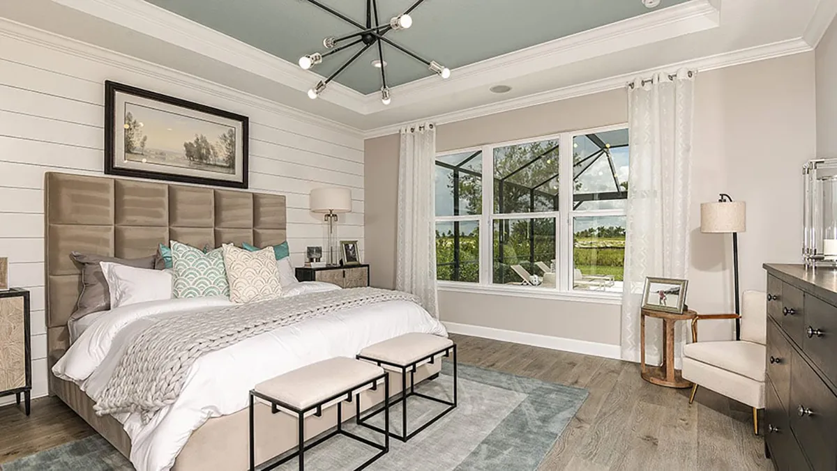 Modern bedroom with a large bed, decorative pillows, two benches at the foot, a dresser, a chair, a lamp, large windows with sheer curtains, and a unique ceiling light fixture. Natural light fills the room.