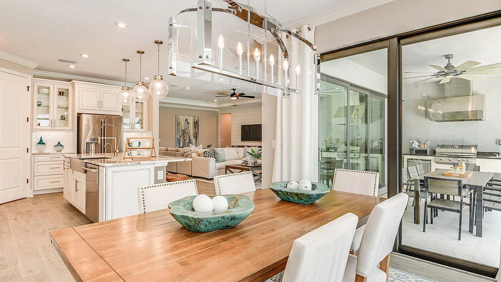 Bright, modern open-concept kitchen, dining, and living area with white cabinetry, pendant lighting, a wooden dining table, green decorative bowls, and large sliding glass doors to an outdoor patio with table and grill.